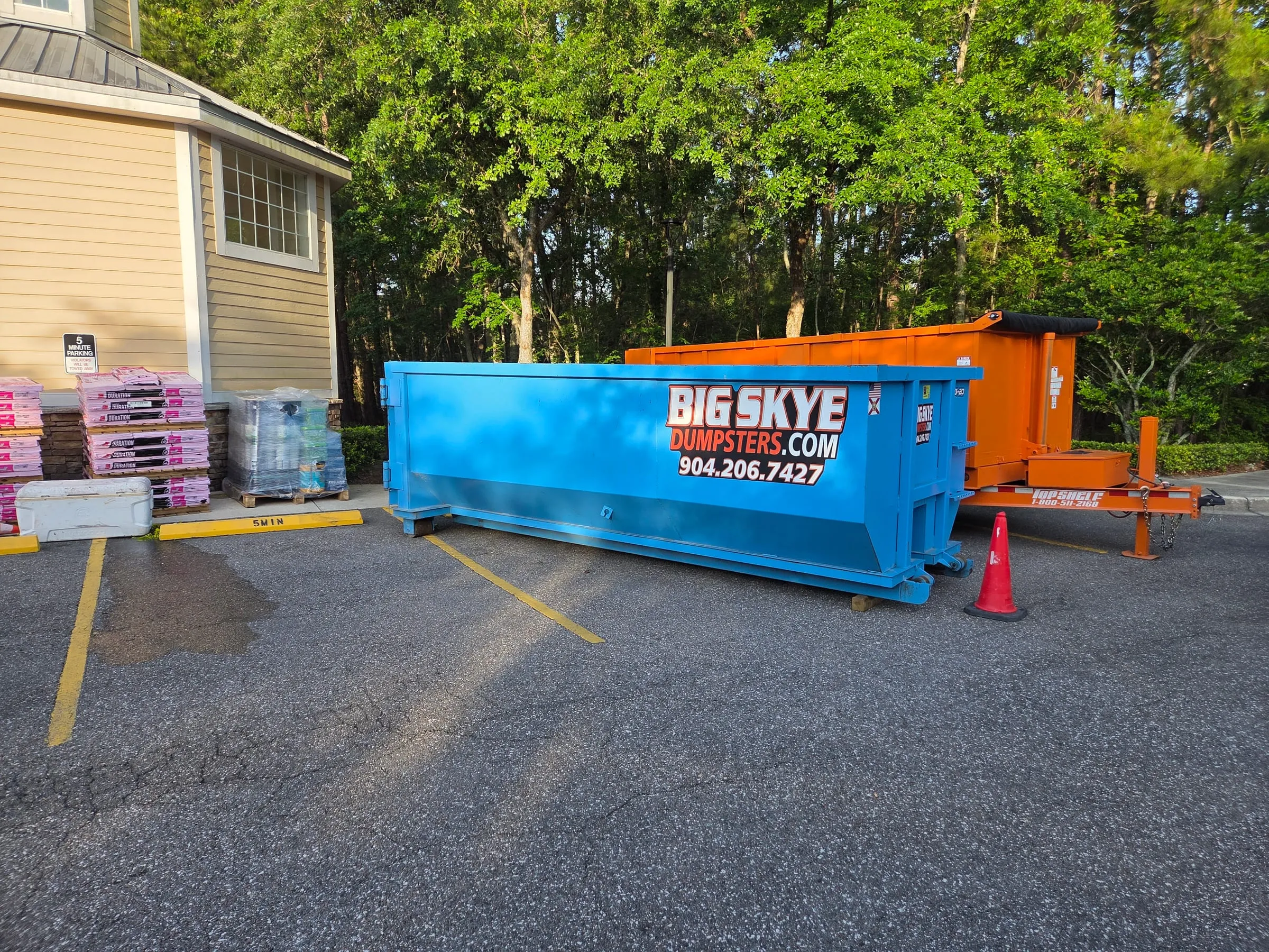 Big Skye roll-off dumpster and an orange dump trailer parked beside pallets of roofing shingles