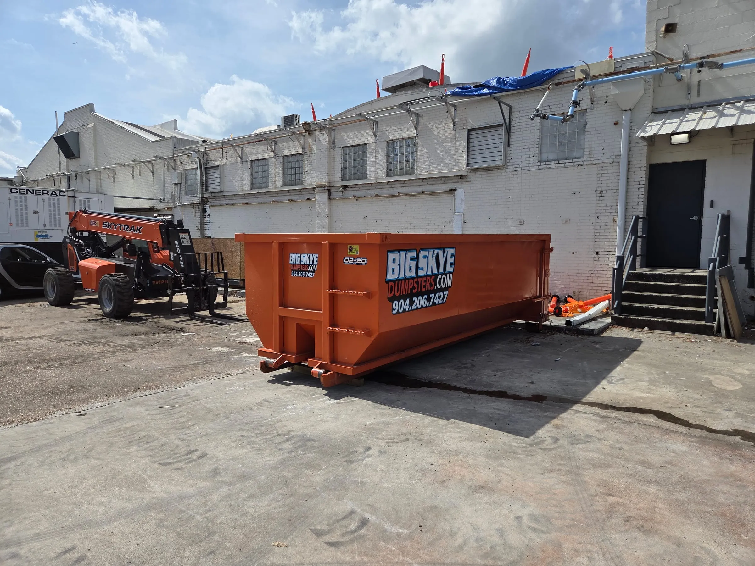 Orange Big Skye roll-off dumpster at a commercial job site next to a telehandler